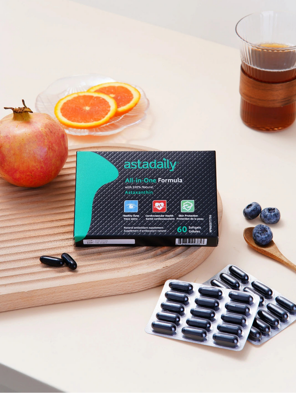 A styled product shot of the astadaily All-in-One Formula supplement box on a light wood board, surrounded by capsules, a pomegranate, orange slices, blueberries, and a glass of iced coffee on a cream-colored table.